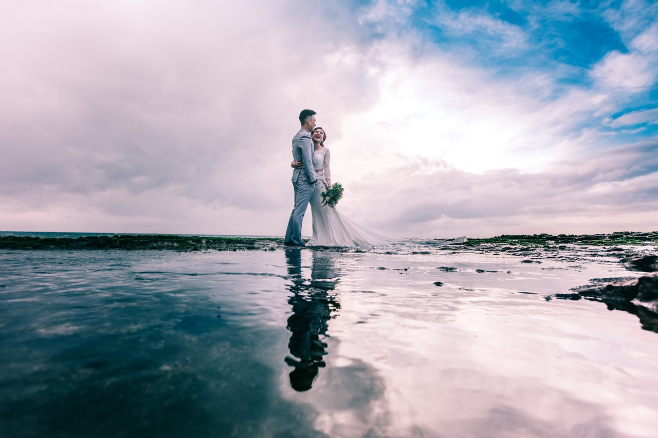 Man in Gray Dress Suit Jacket Embraces Woman Wearing Wedding Gown near the sea