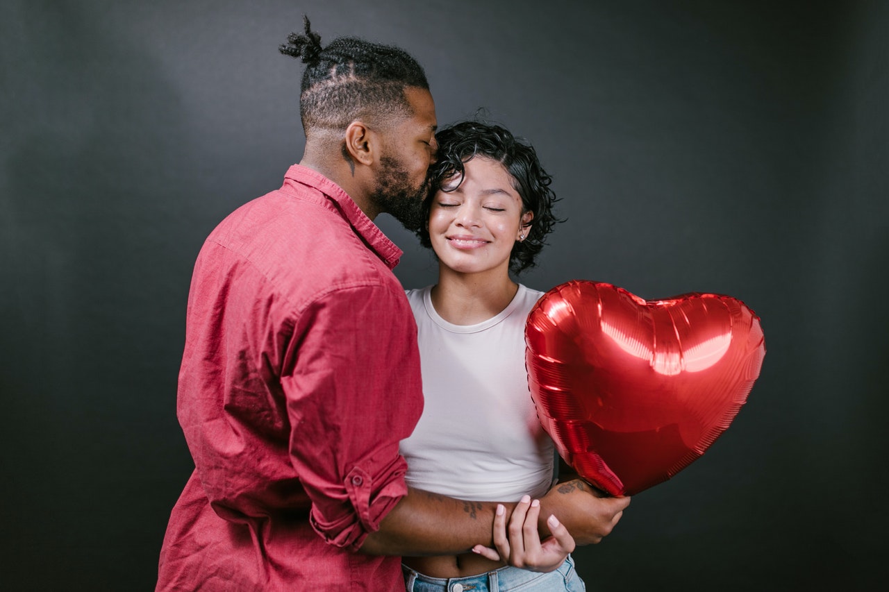 Man Kissing His Woman While Holding a Red Heart Shaped Balloon