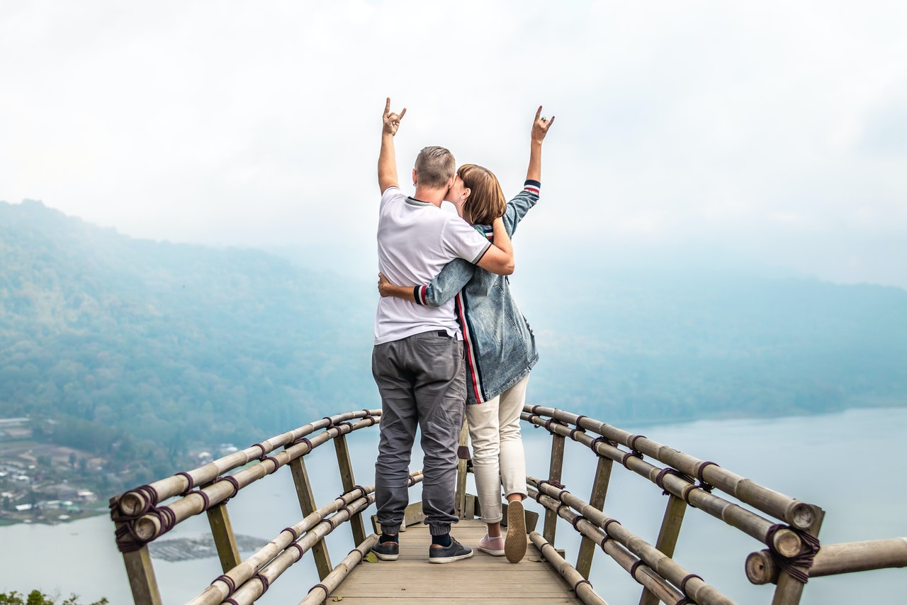 Couple Hugging Each Other on Hand Rails With Arms Wide Open Facing the Mountains and Clouds with their hands up
