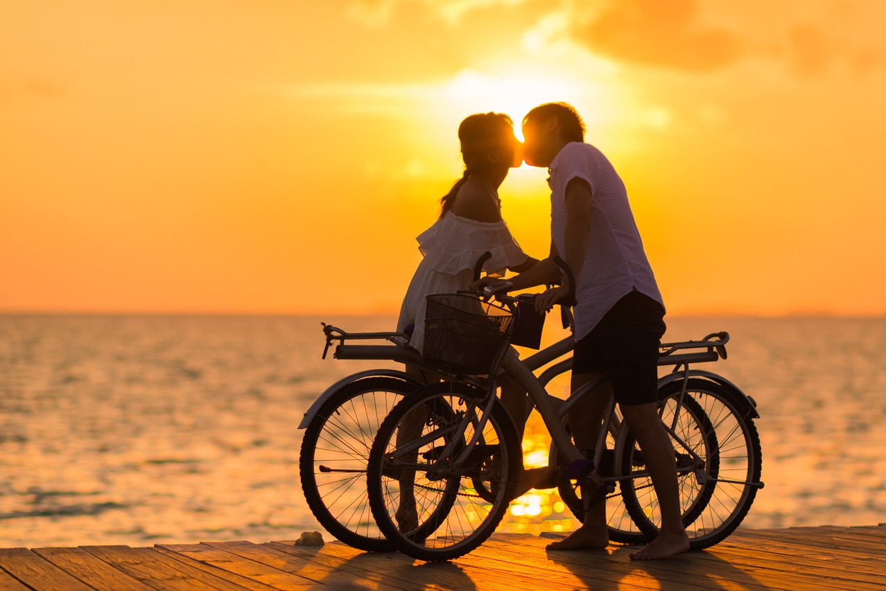 Man Wearing White T-shirt Kissing a Woman While Holding Bicycle on River Dock during Sunset