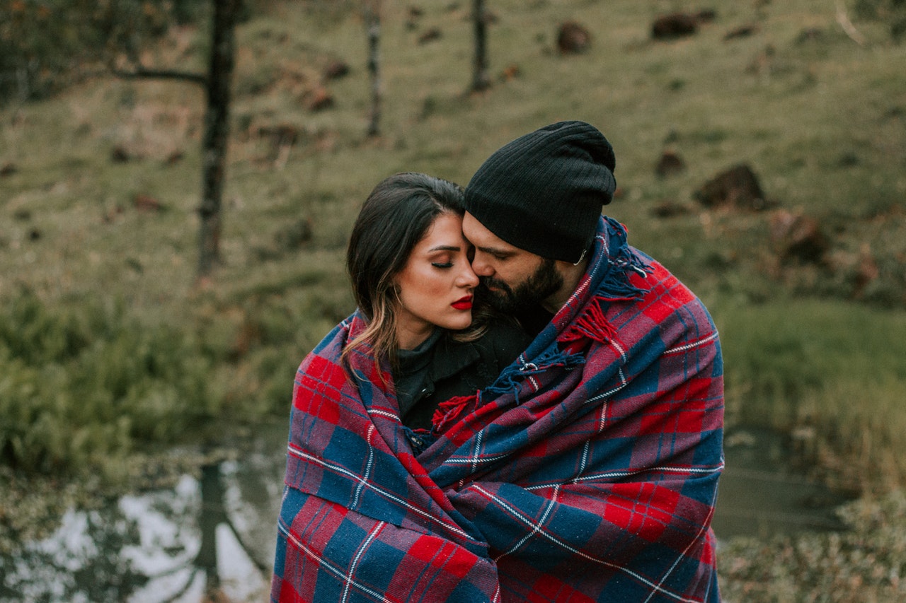 A Couple Hugging While Covered in Red and Black Blanket Outdoors