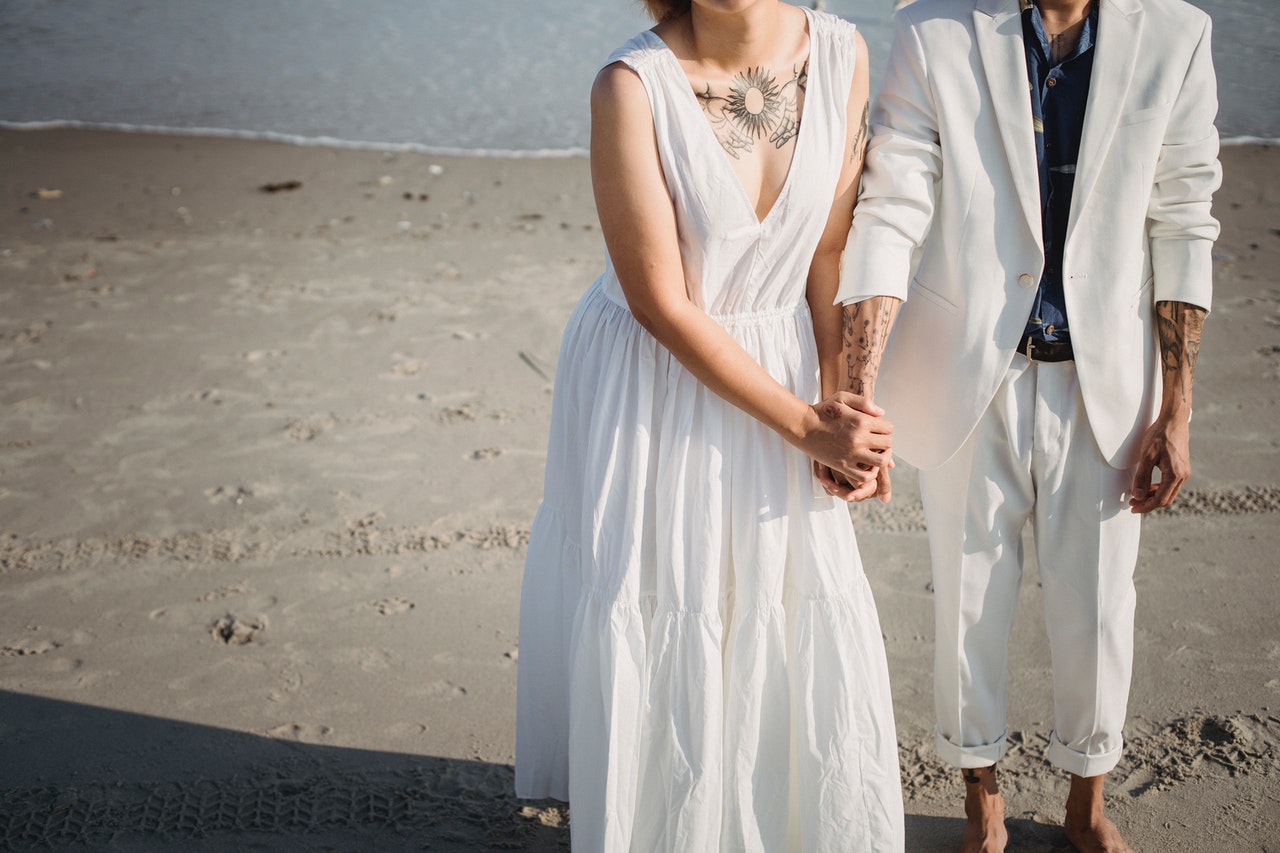 A Woman in White Sleeveless Dress And A Man In White Suit And Pants Are Standing on Beach Sand