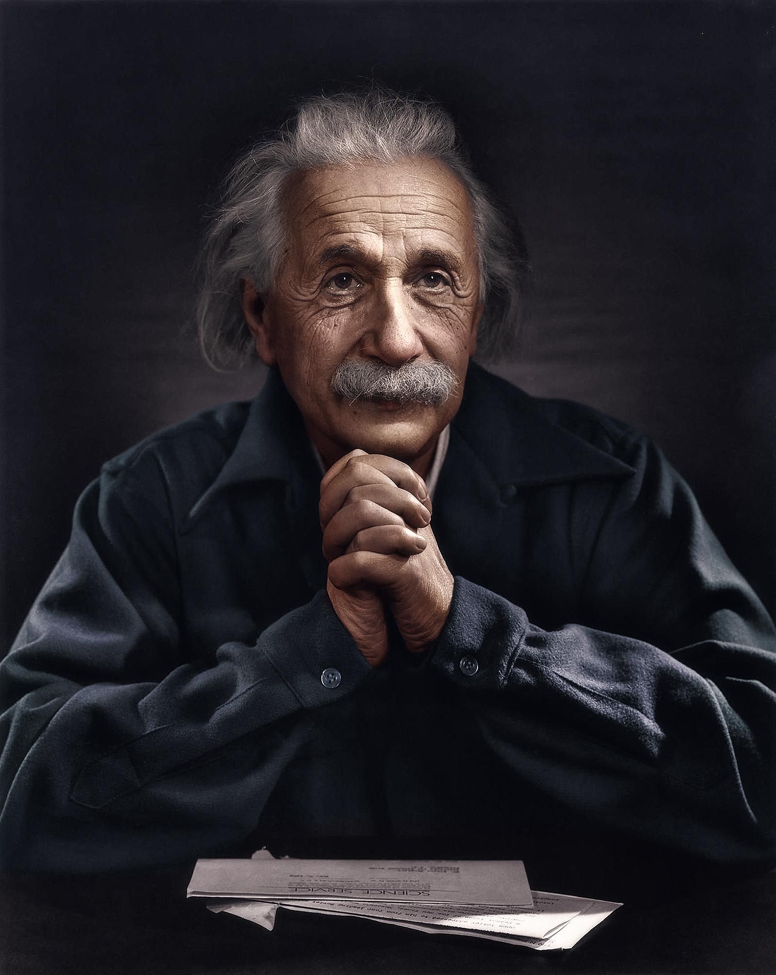 Alber Einstein sitting with his hands clasped together with papers in front of him