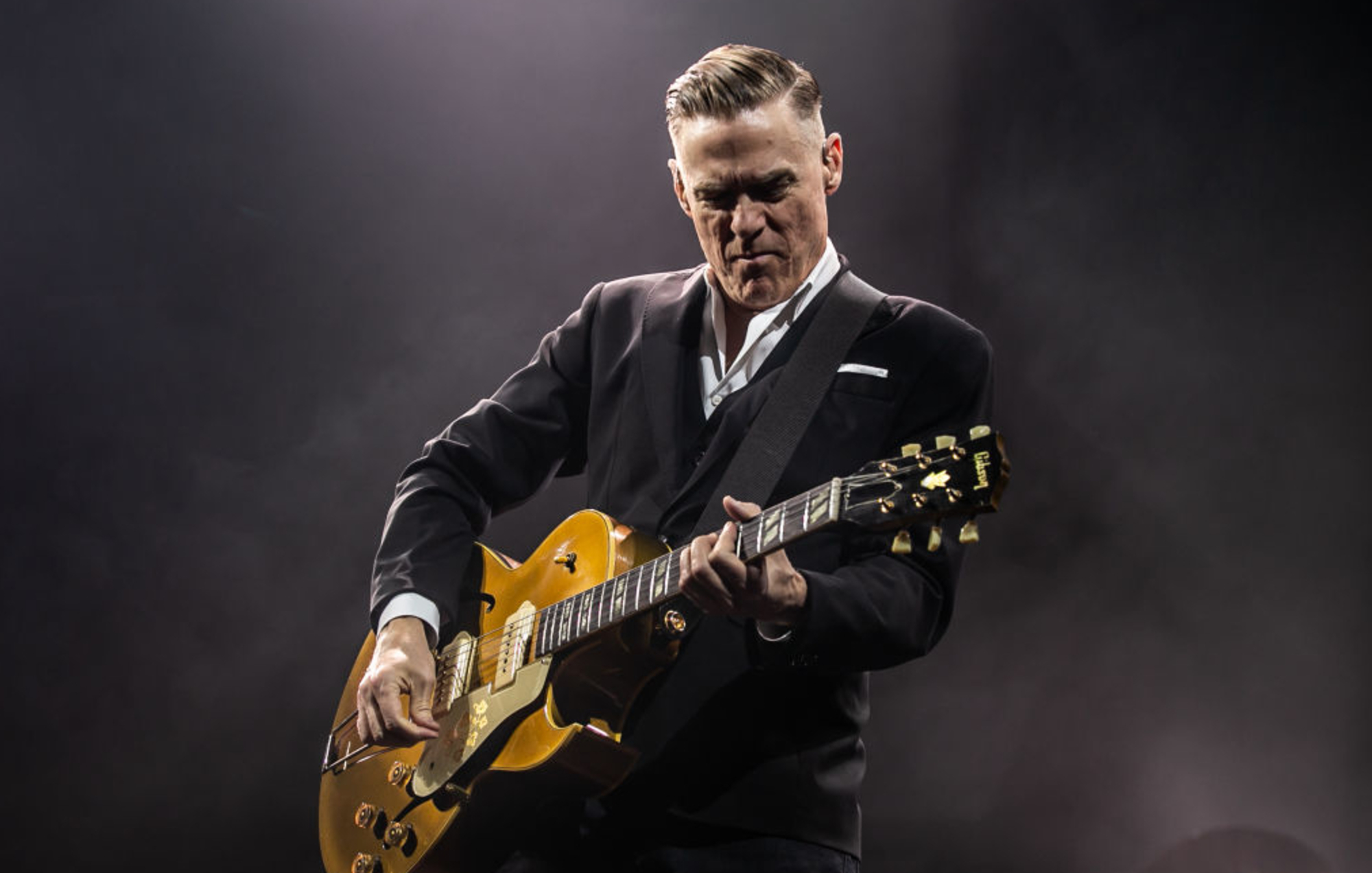 Bryan Adams wearing a black suit while playing a guitar