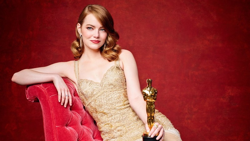 Emma Stone holding a trophy while sitting on a red couch