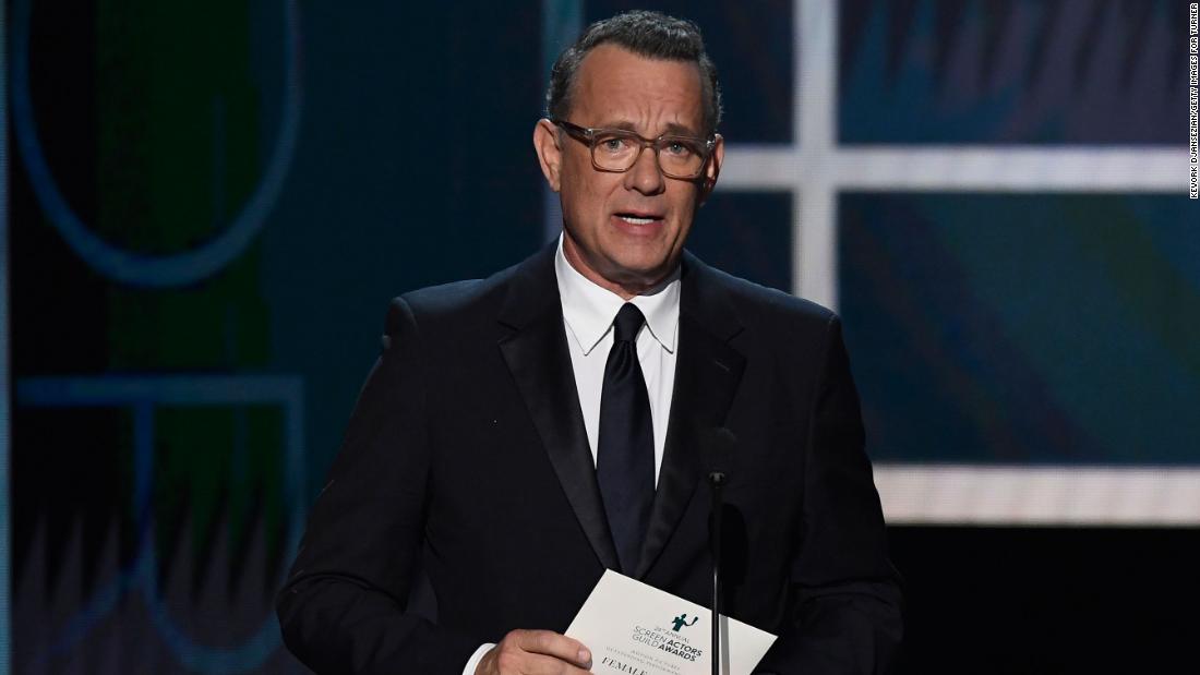 Tom hanks wearing a black suit and holding a paper while in front of a microphone