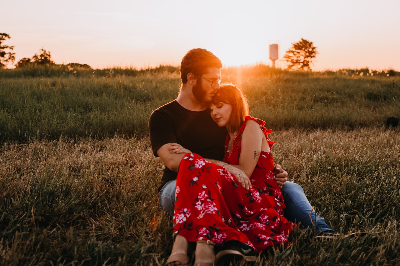 Man in Black Crew Neck T-shirt Sitting on Grass Field Beside A Woman in Red Floral Dress