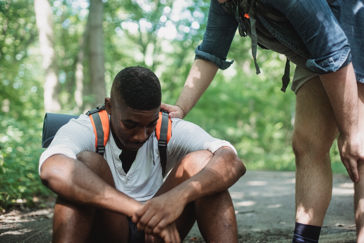 Unhappy black hiker sitting on ground in forest with another person's hand on his shoulder
