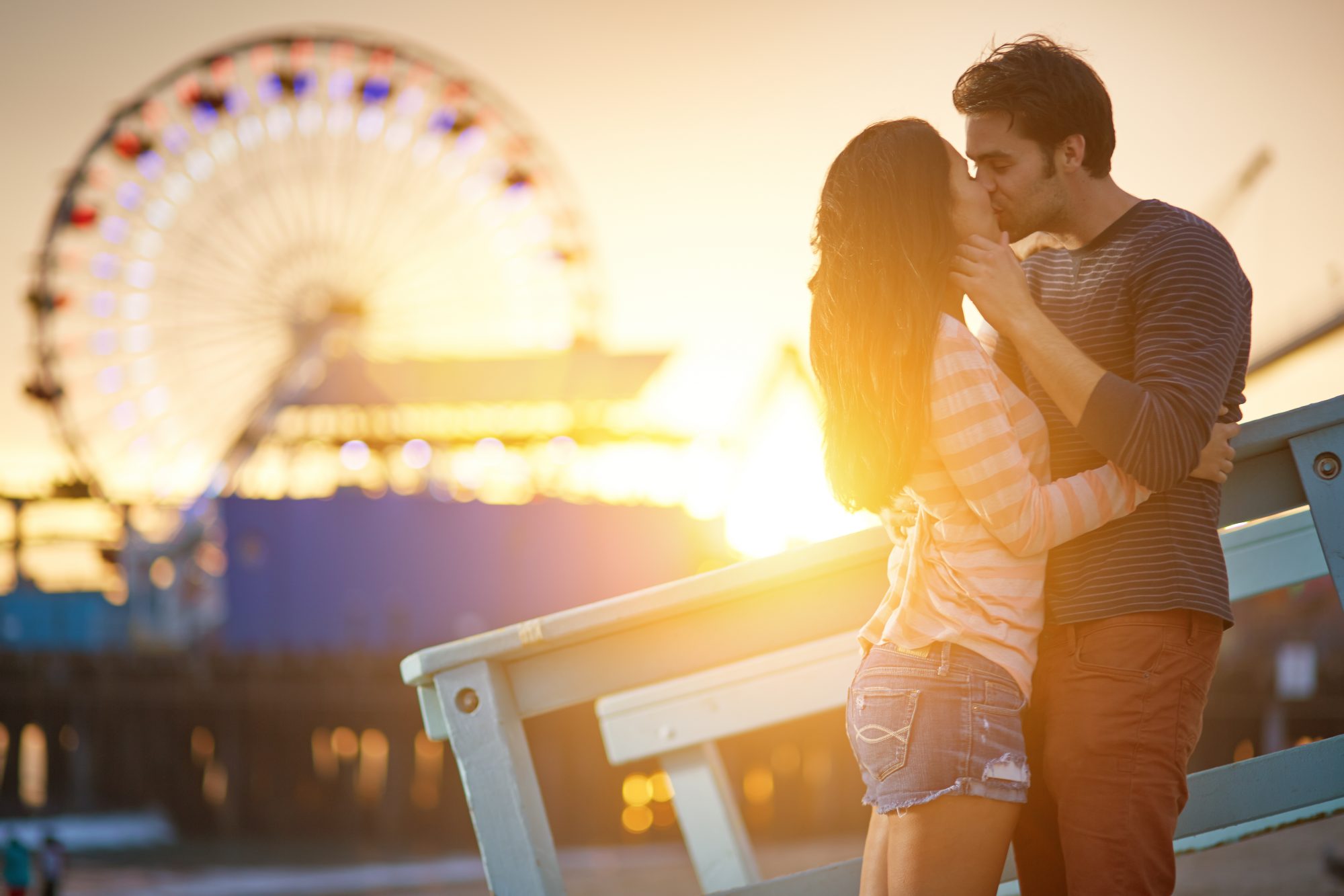 A man and woman kissing at a park with a Ferris wheel behind of them