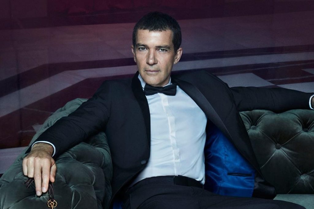 Antonio banderas sitting on a big sofa while wearing a black coat and black bow tie
