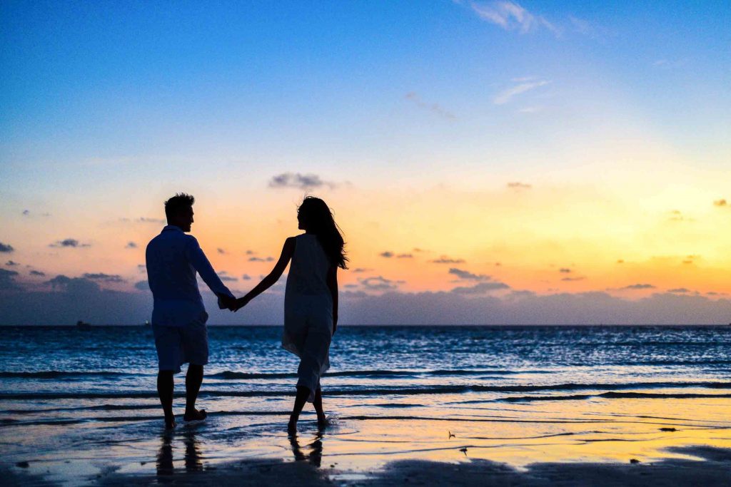 Man and woman holding hands walking on seashore during sunset