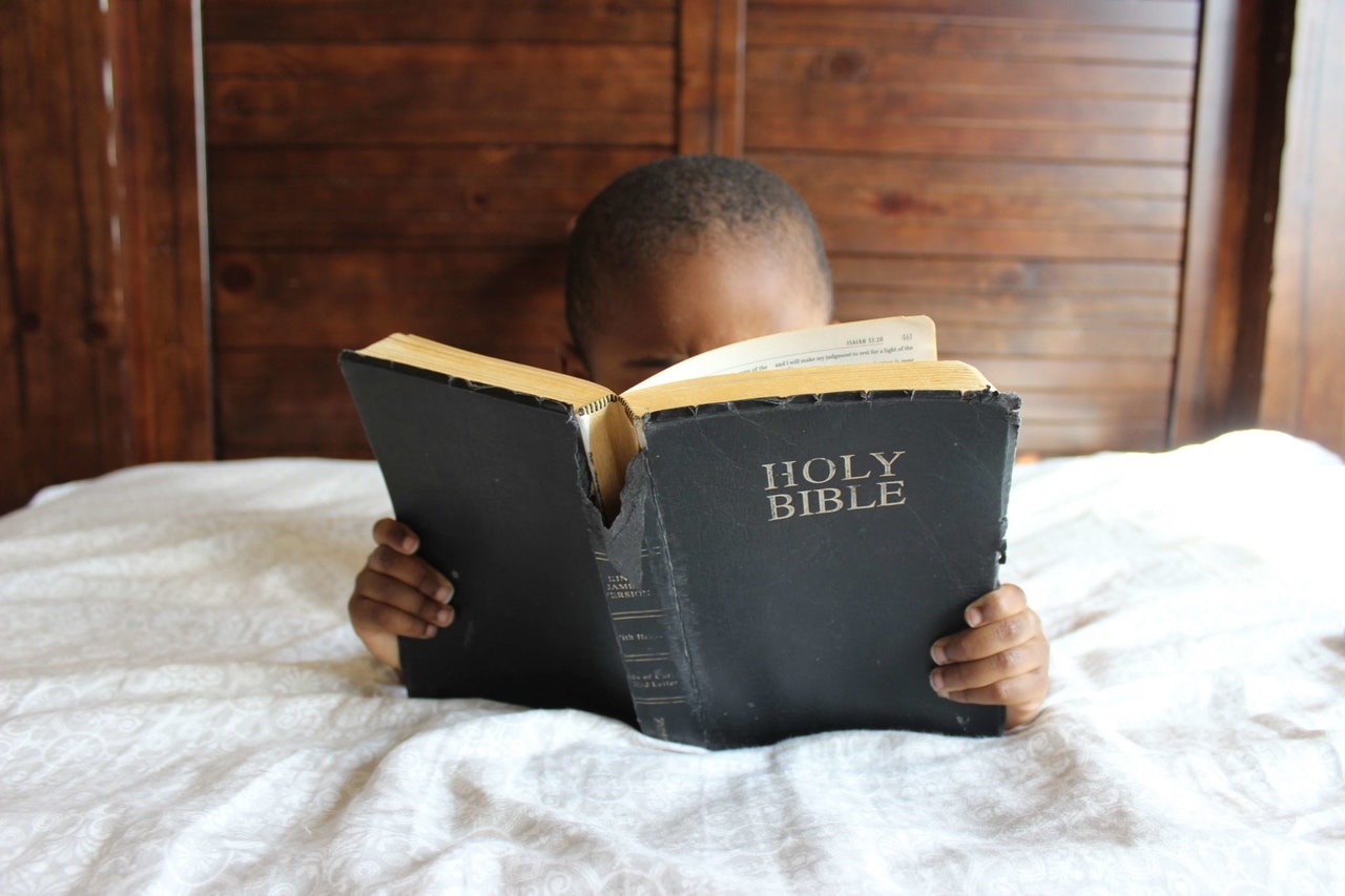 Child Reading Holy Bible On The Bed