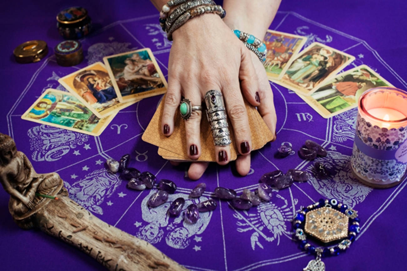 A psychic holding a tarot card having a candle, beads and pendulum on the table