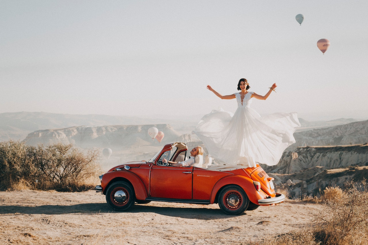 A bride standing on a red volkswagen beetle while the groom is on the driver's seat