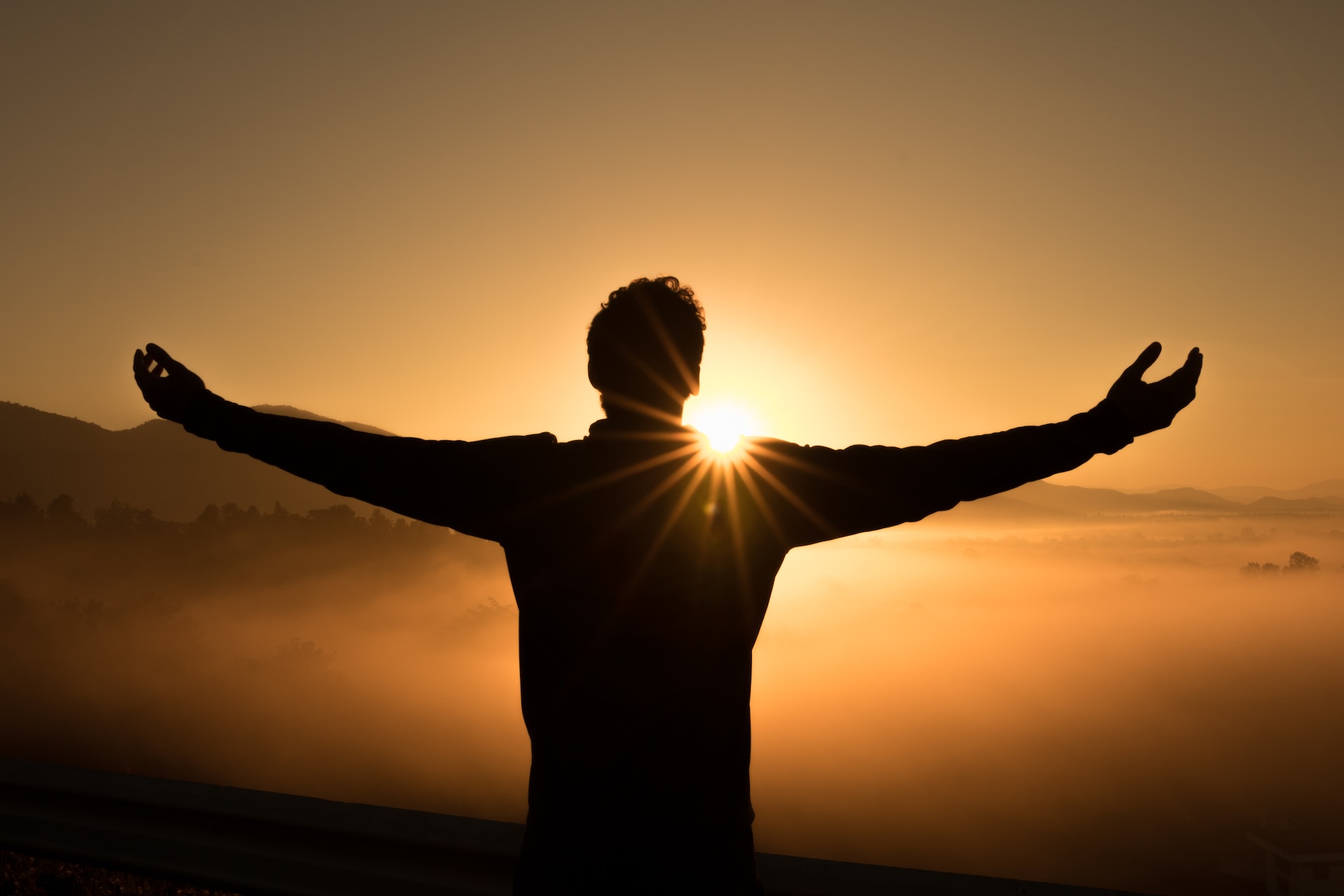 Silhouette of a man with arms wide open and facing a sunrise, with sunlight peeking through his left shoulder