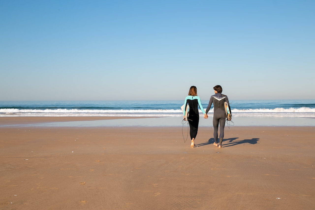 Couple in full body swimwear walking on shore with surfboards on their arms