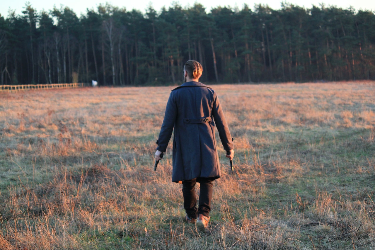 A Man In Black Coat Walking In A Field While Holding A Gun