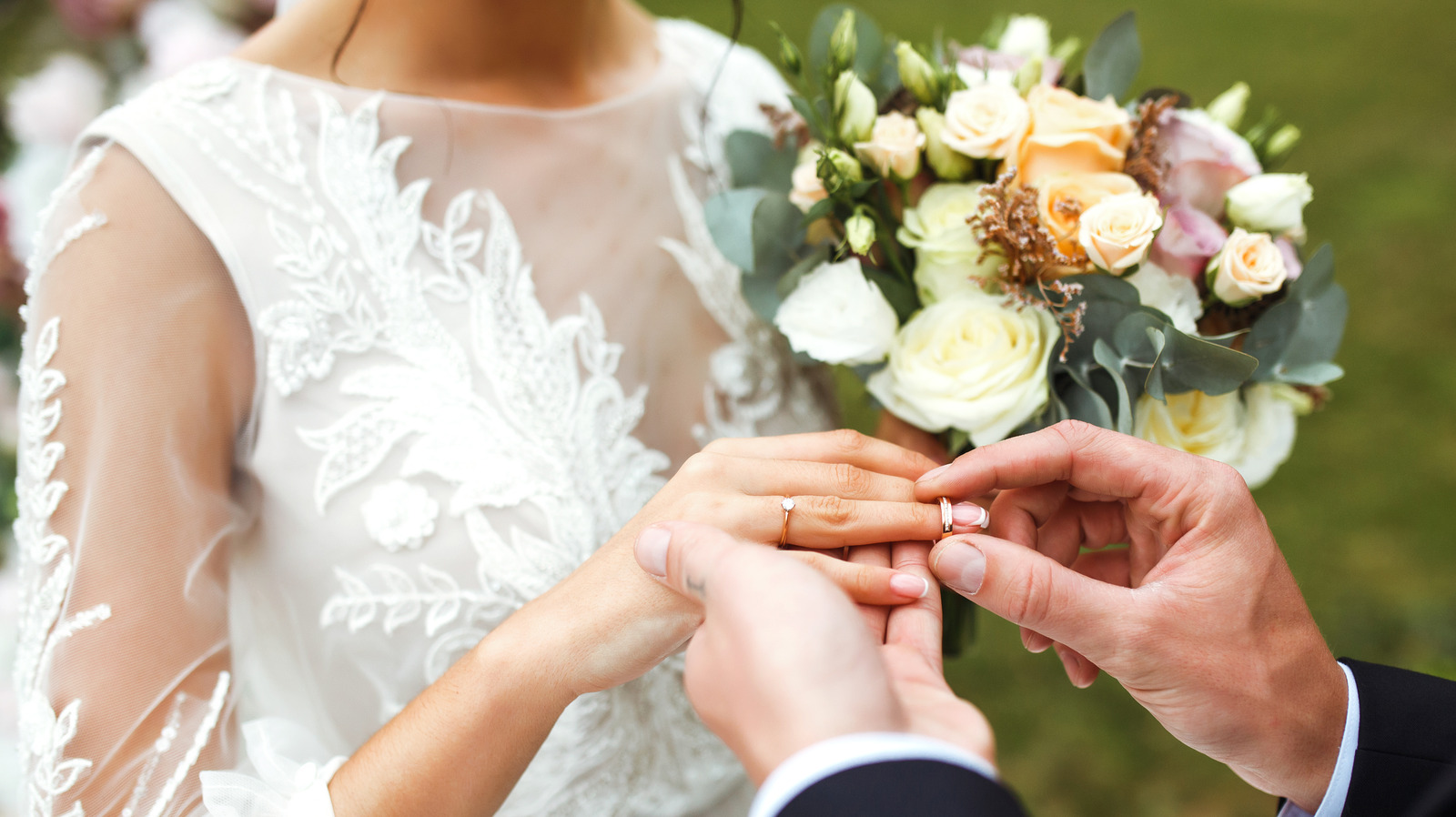 A man putting a ring on a girl's hand in a weading dress and holding a bouquet of flowers