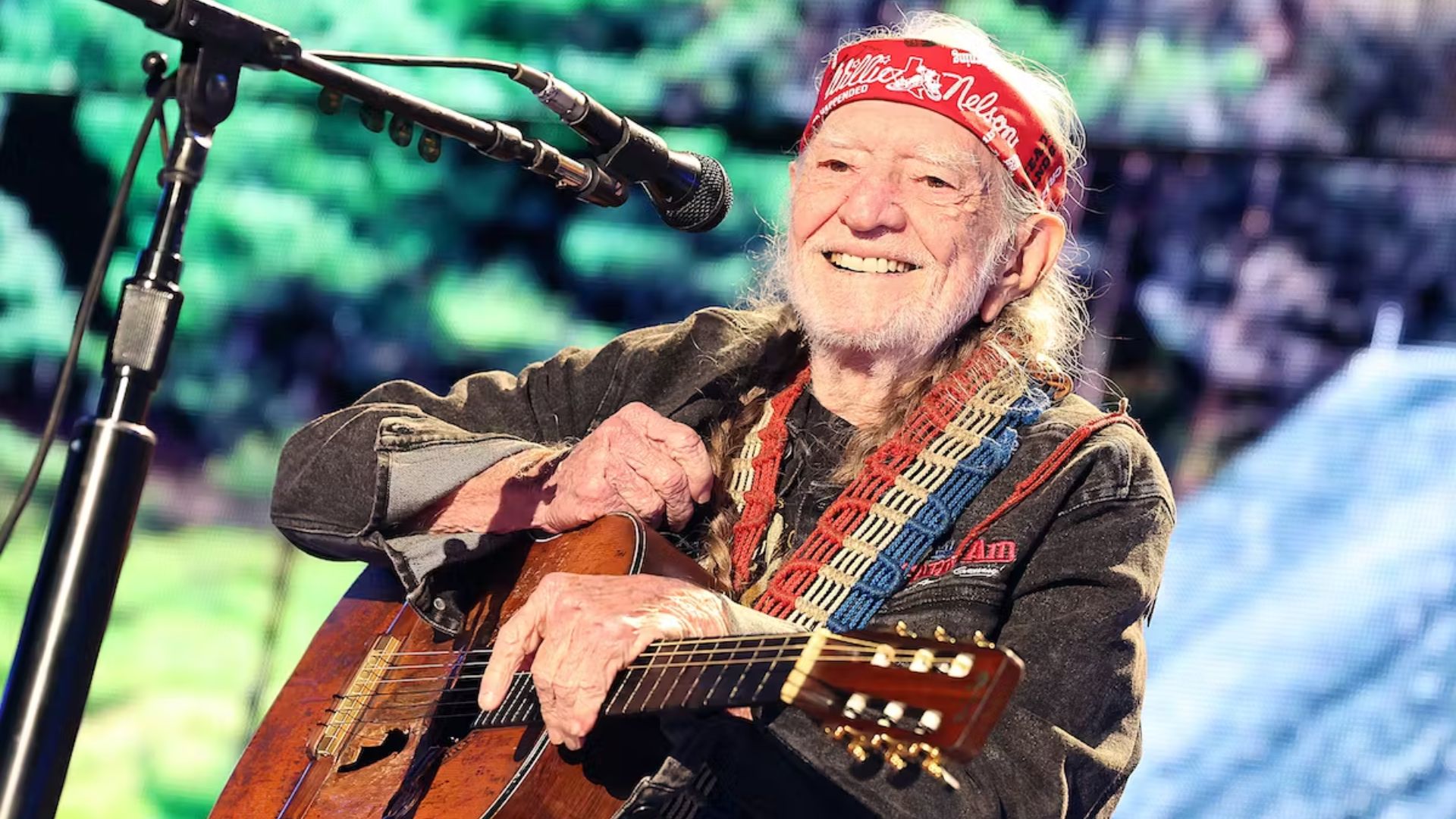 Willie Nelson Holding A Guitar In Front Of A Mic