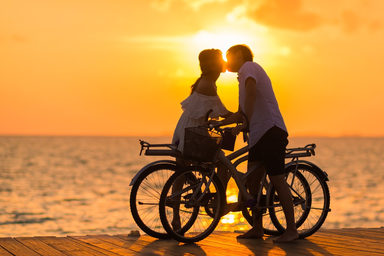 Man Wearing White T-shirt Kissing a Woman While Holding Bicycle on a Dock during Sunset