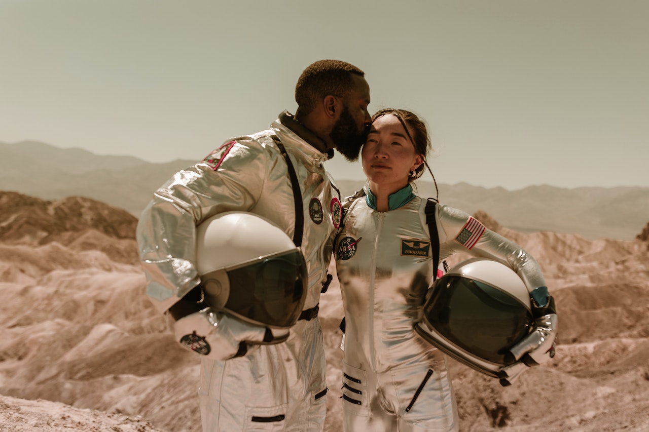 A Couple Wearing Space Suit, Holding Helmets Where The Man Kisses Woman's Temple