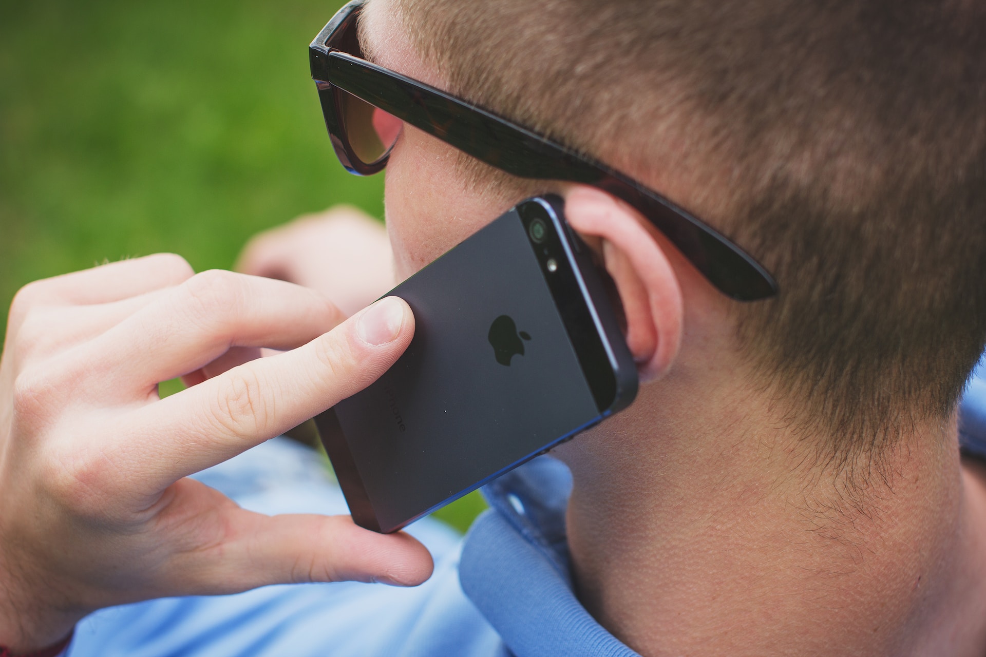 A man in dark shades and light blue collared shirt calling someone on his black iPhone 5s