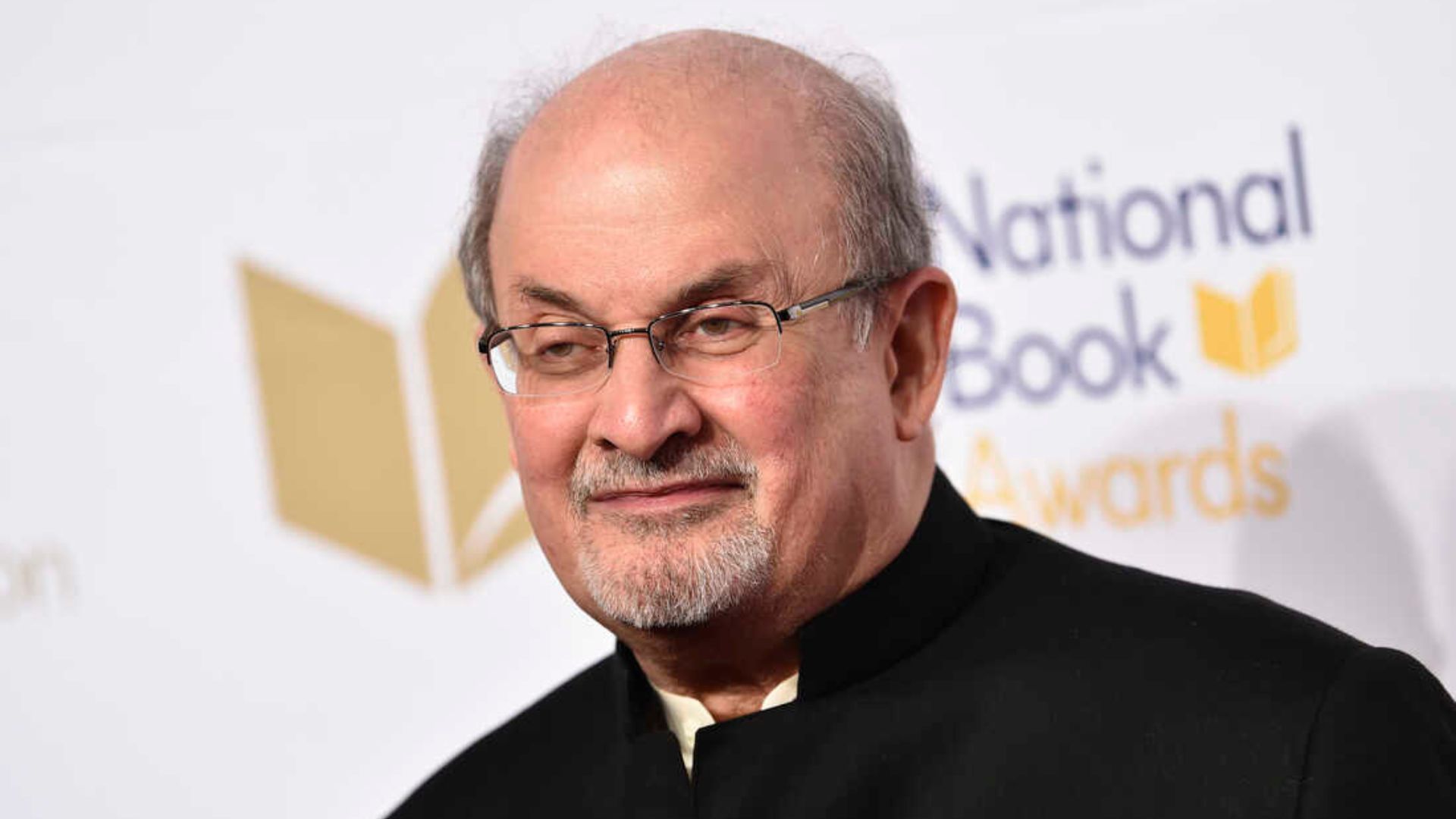 Salman Rushdie Smiling And Standing In Front Of A White Poster