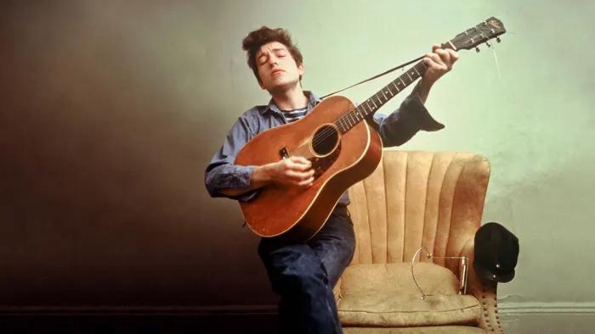 Bob Dylan Playing a Guitar while Sitting on the Arm of a Chair