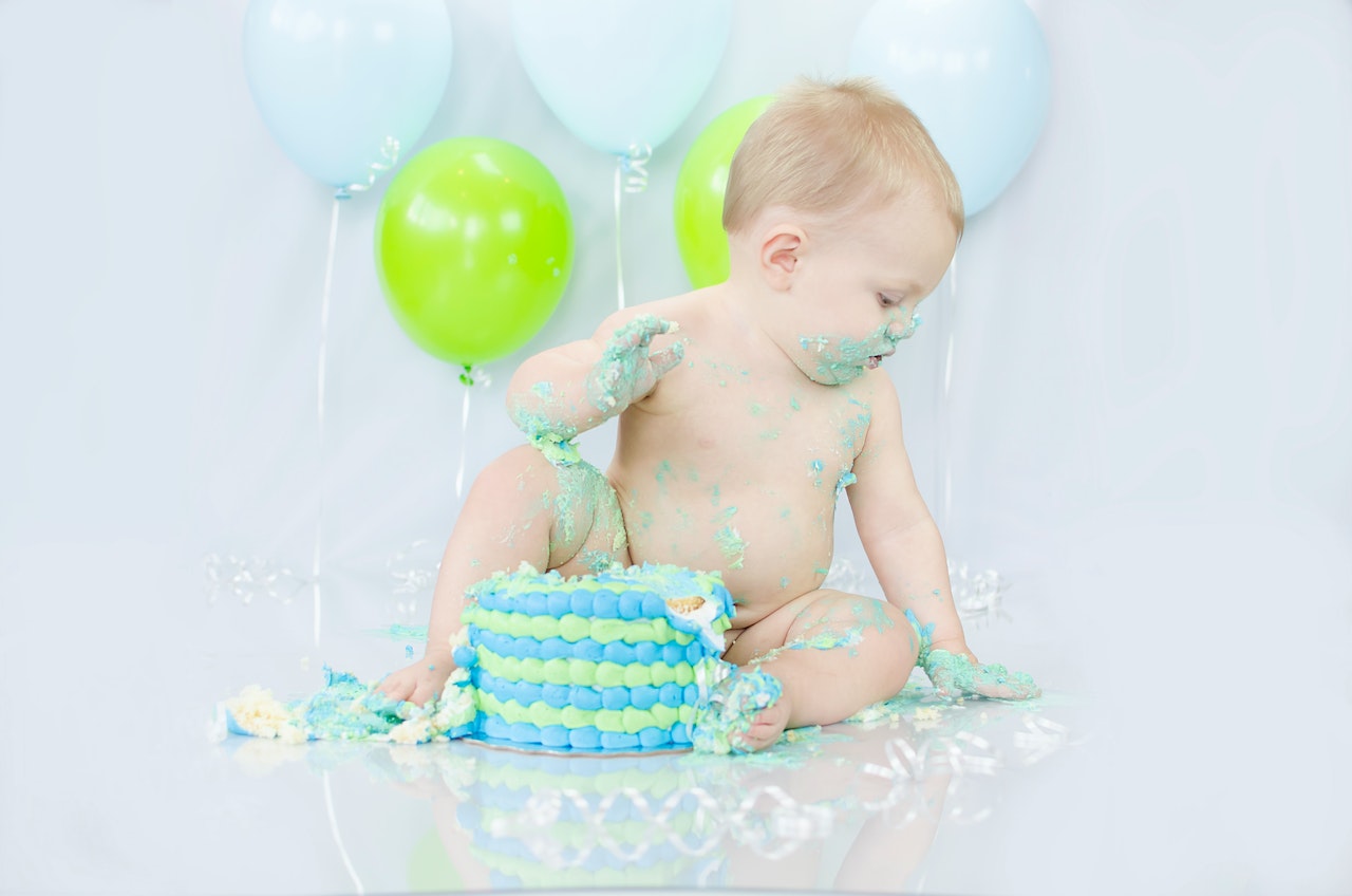 A Baby Sitting Beside Blue and Green Cake and Balloons