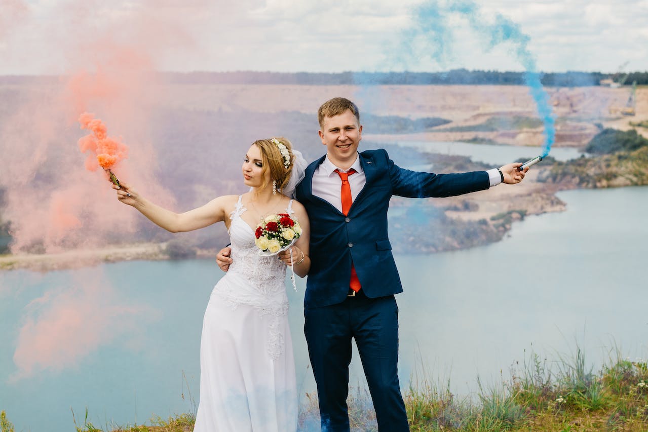 Bride And Groom Holding Smoke Bombs Near Body Of Water